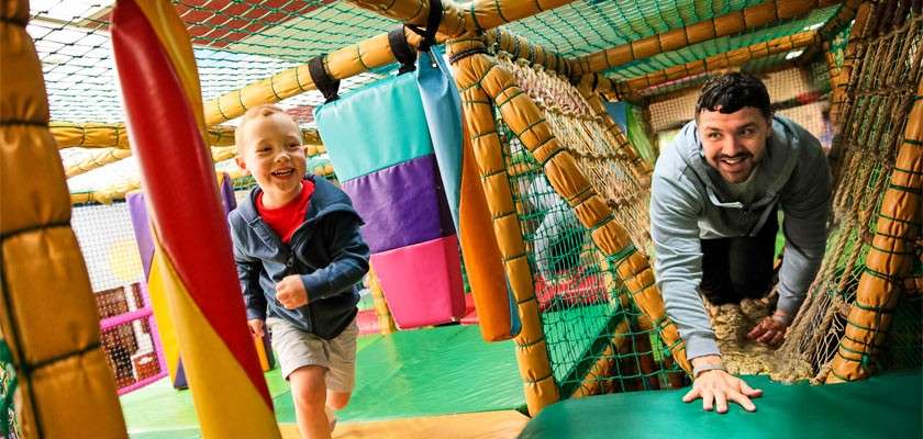 Child runs while a man crawls through a netted tunnel in an indoor soft-play structure, surrounded by padded beams, colorful foam blocks, mesh walls, and bright mats.