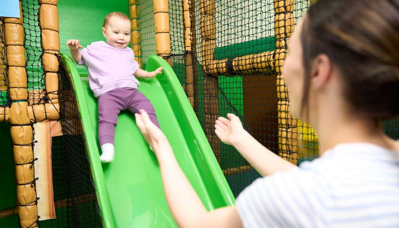 Toddler slides down a bright green slide, smiling; adult with outstretched arms waits to catch; surrounded by netted, padded play structures in an indoor playground.