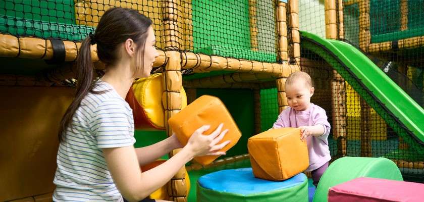 Adult and toddler stack large soft foam cubes on padded platforms amid netted climbing structures and a green slide in an indoor playground.