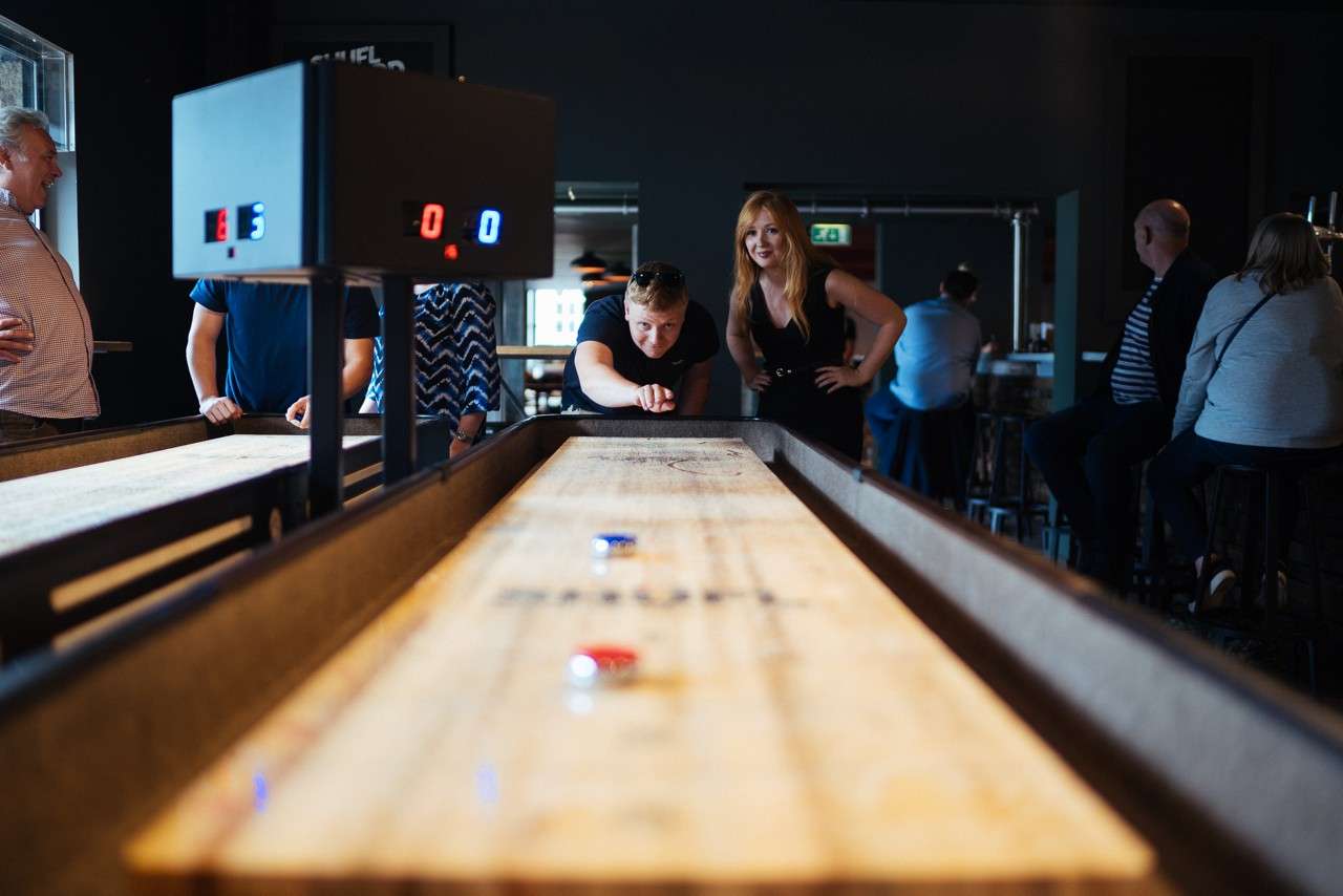 Player pushes a puck down a long wooden shuffleboard table as spectators watch in a bar. Electronic scoreboard displays: 6, 0, 0. People stand and sit around the table, observing.