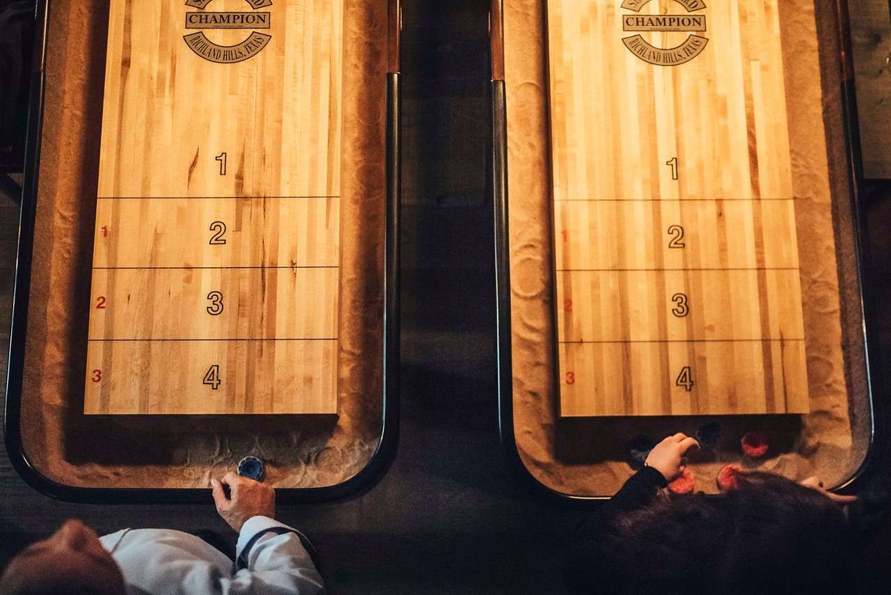 Two shuffleboard tables host players readying pucks in a dim indoor game area with sanded gutters. Text: CHAMPION; RICHLAND HILLS, TEXAS; 1 2 3 4; 1 2 3; CHAMPION; RICHLAND HILLS, TEXAS; 1 2 3 4; 1 2 3.