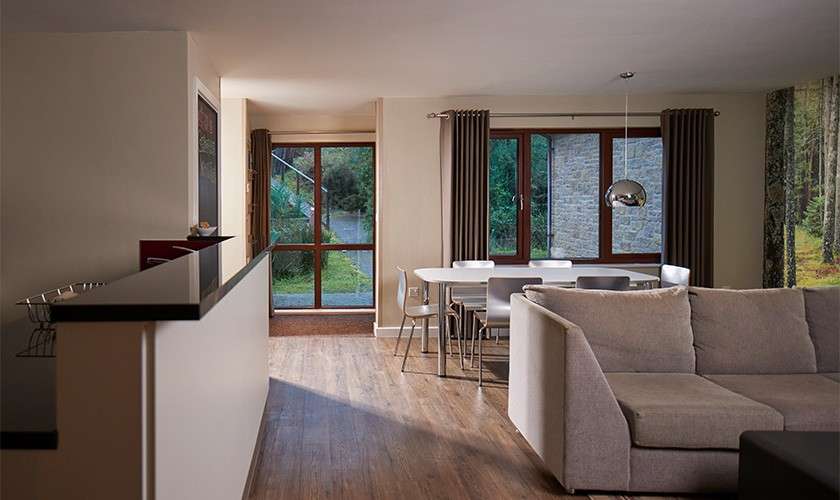 A gray sectional sofa faces an open-plan kitchen; a dining table sits under a metallic pendant; large windows admit natural light, revealing greenery and a stone wall outside.