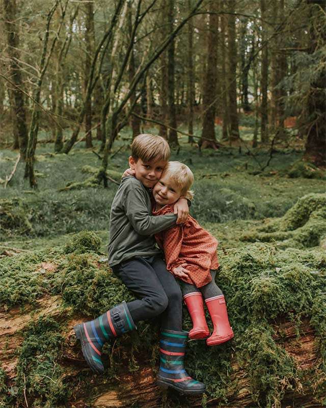 Two children sit hugging on a moss-covered fallen log, wearing colorful rain boots, surrounded by dense, green forest with tall trees and soft ground.