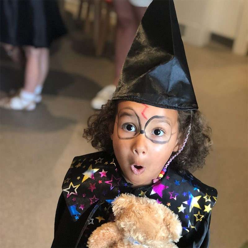 Child in wizard costume gasps with wide eyes, holding a teddy bear. Indoors, other people blurred in background; starry cape, pointed hat, face painted with glasses and lightning-bolt scar.