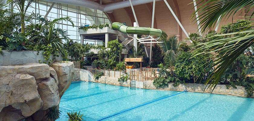 Indoor swimming pool glistens; a blue lane line stretches across while a green tube waterslide curves overhead. Tropical plants, rock formations, and a glass roof create a lush resort setting.