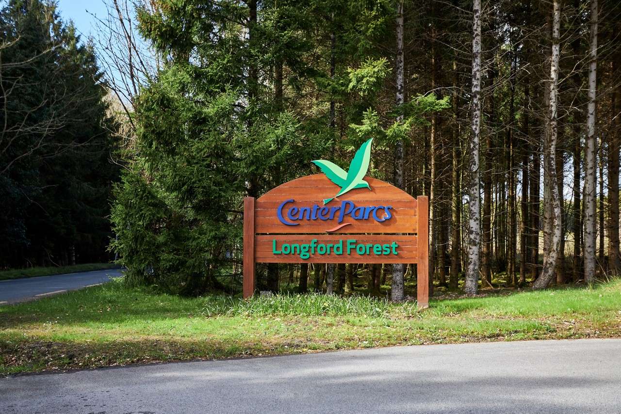 Wooden entrance sign displays resort name beside a road at a forest edge. Text: “Center Parcs” and “Longford Forest.”