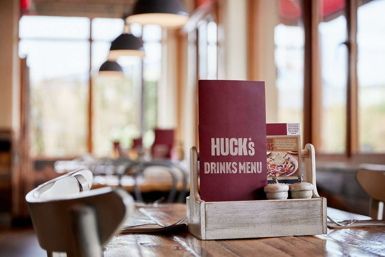 Drinks menu stands upright in a wooden caddy with salt and pepper shakers on a polished restaurant table; blurred chairs, windows, and pendant lights in background. Text: HUCK's DRINKS MENU.