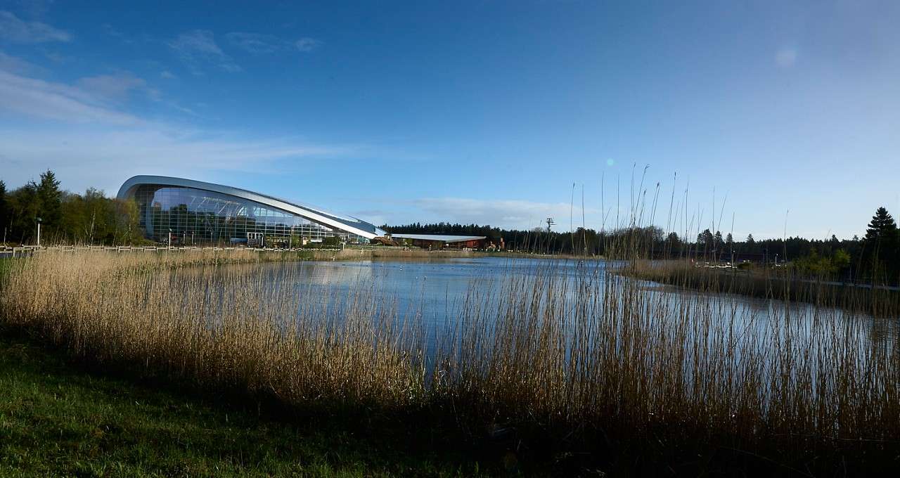 Curved, glass-walled building overlooks a calm lake, reflecting light. Tall reeds line the water’s edge, with trees and a distant treeline under a clear blue sky.