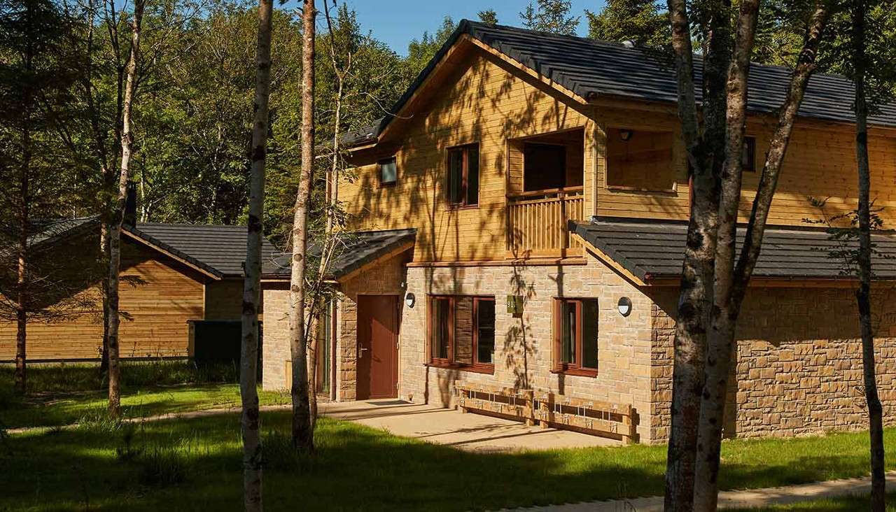 Two-story lodge stands with stone lower walls, wooden upper level and small balcony, facing a paved path, surrounded by tall trees and sunlit grass in a quiet forest clearing.