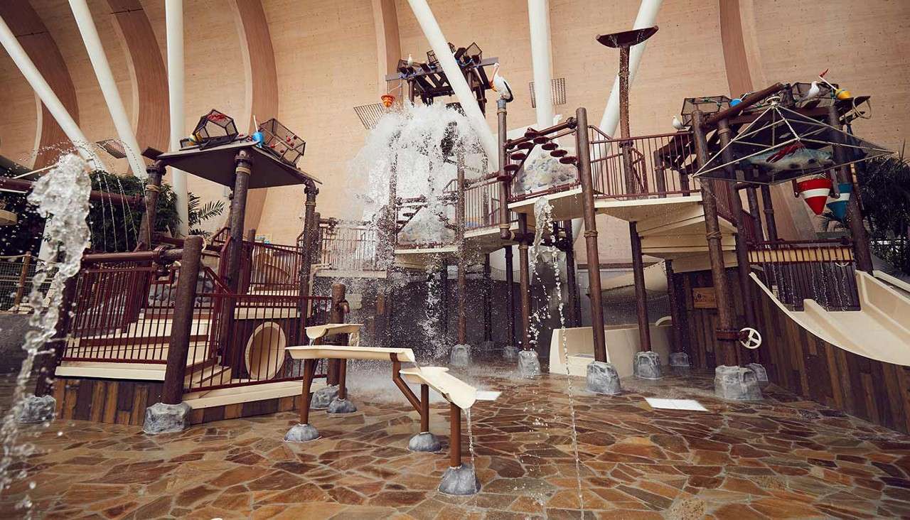 Water-play structure splashes; a giant tipping bucket dumps water while sprays arc, slides and stairways surround. Indoors with high arched beams, rock-pattern floor, nautical-themed features.