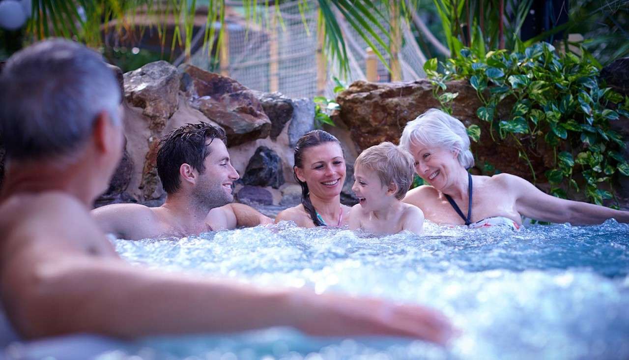 Five people lounge and laugh in a bubbling hot tub, chatting and holding a child, surrounded by rocks, tropical plants, and a netted play structure in the background.