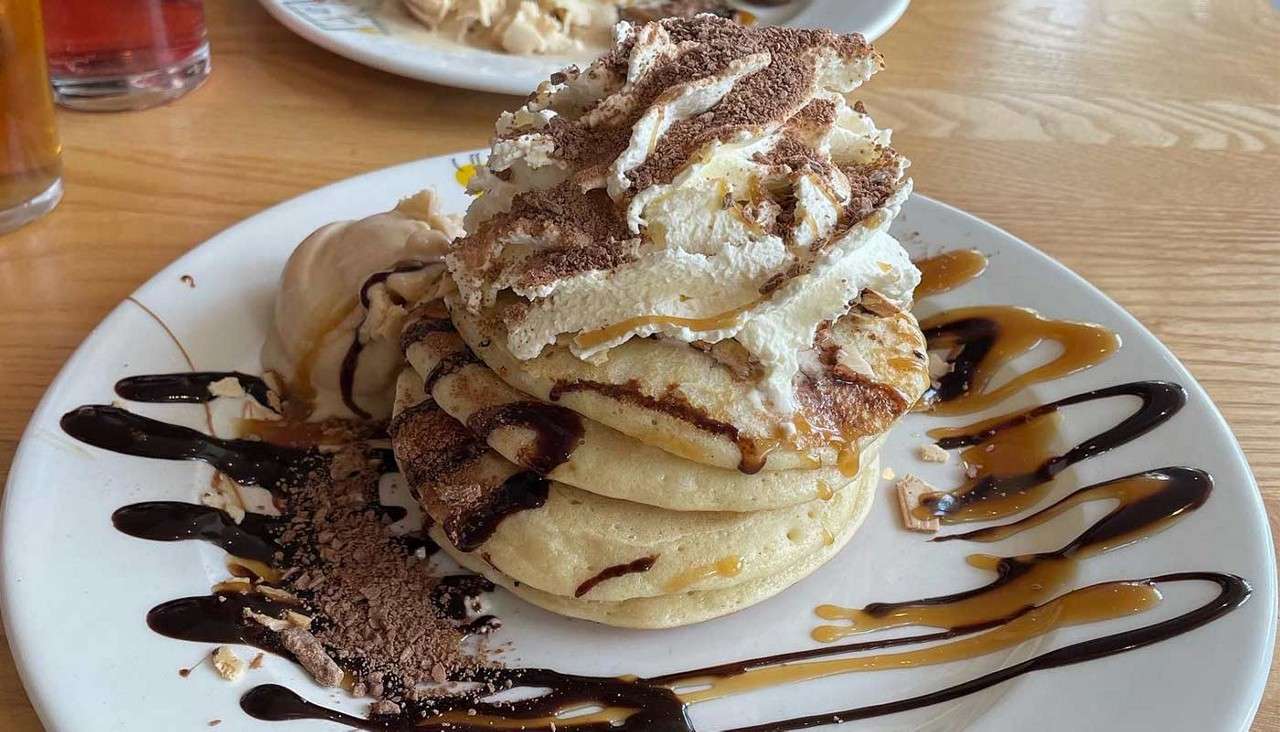Stacked pancakes topped with whipped cream and cocoa crumble, drizzled with chocolate and caramel; a scoop of ice cream melts beside them on a white plate on a wooden table.