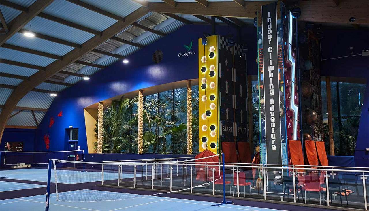 Climbing walls stand ready beside badminton courts, inside a blue-roofed sports hall with large windows and tropical plants. Visible text: Indoor Climbing Adventure; START; START; Center Parcs; nature walks.