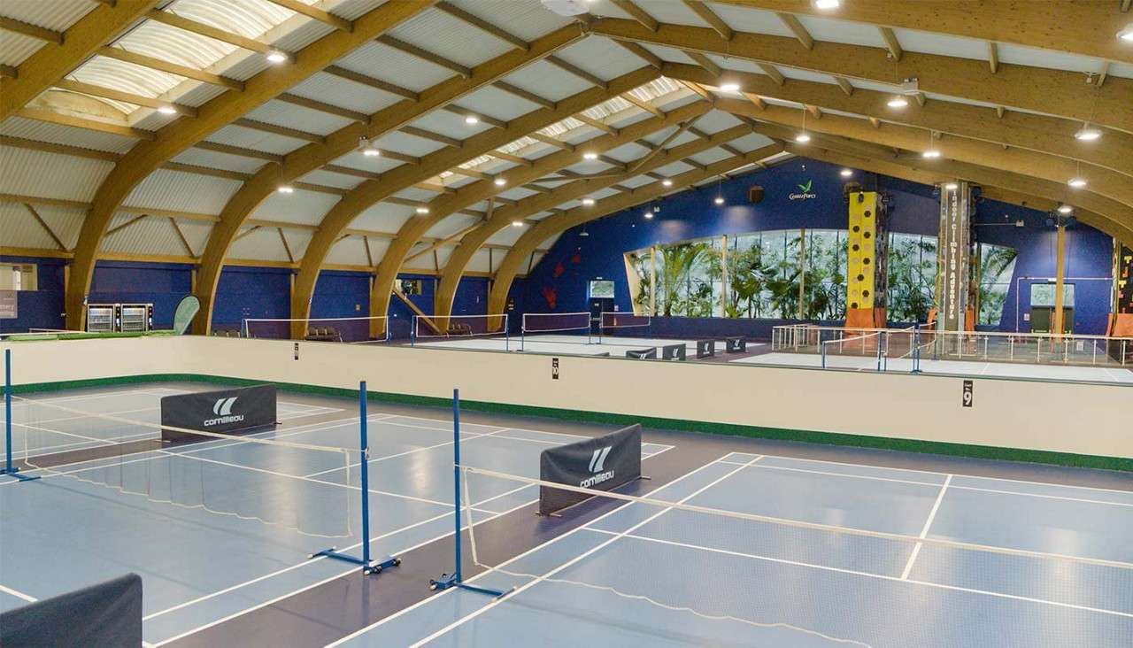 Multi-sport courts sit unoccupied, divided by low nets and cornilleau barriers, within a timber-arched indoor arena featuring blue walls, climbing structures, and large windows. Text: cornilleau; GameZone.