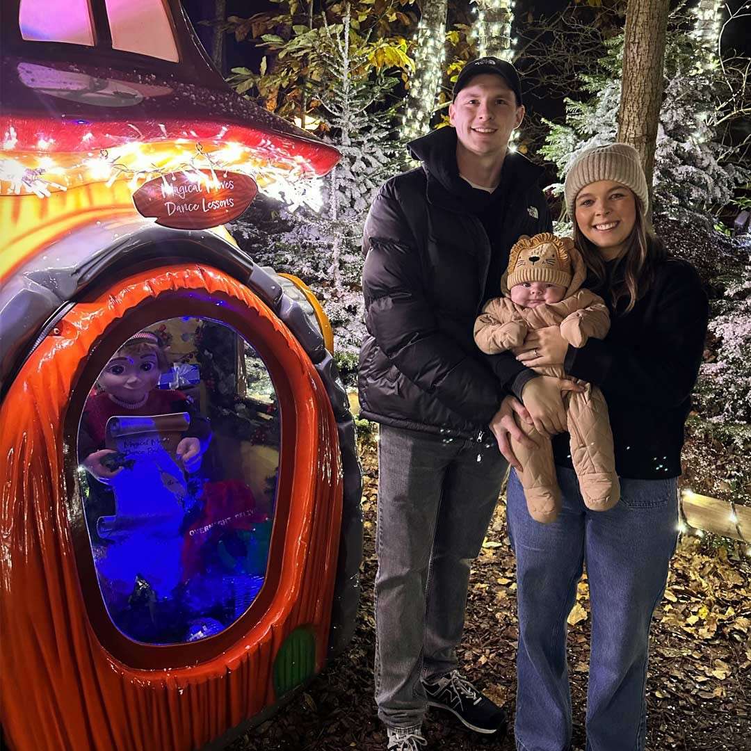 Two adults hold a bundled baby, posing beside a whimsical orange display window with an elf figure; the scene is a snowy, tree-lit holiday setting at night. Text: “Magical Moves Dance Lessons.”