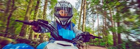 Child on a small ATV rides forward, wearing a full-face helmet and blue vest; hands gripping handlebars; surrounding forest blurred with motion, indicating speed along a wooded trail.