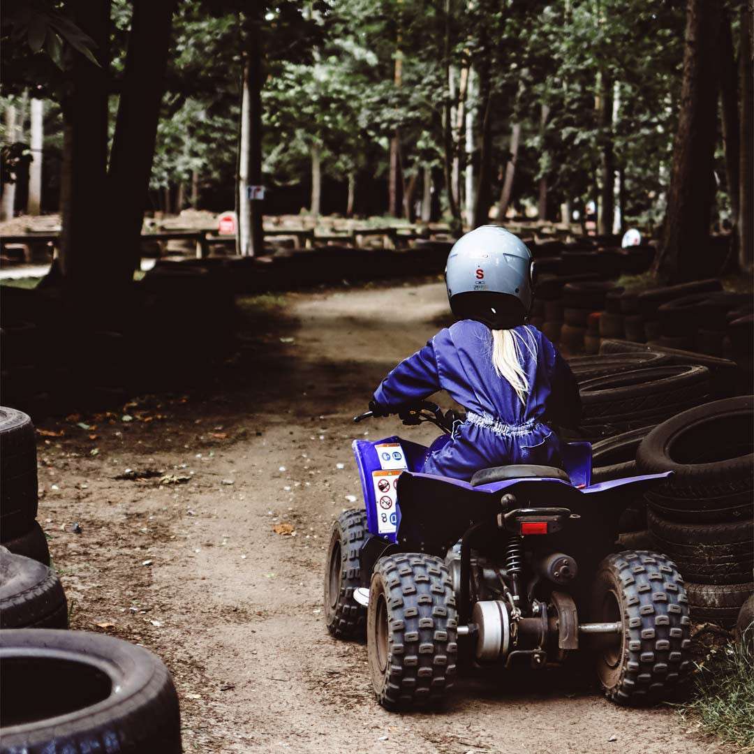 Rider steers a small quad bike along a dirt path, wearing a helmet labeled “S,” amid stacked tires on a forested track.