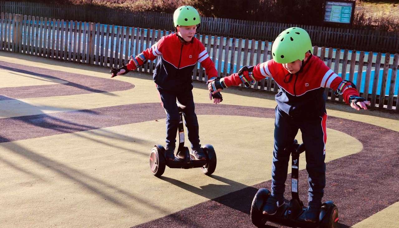 Two children ride self-balancing scooters, arms outstretched for balance, wearing green helmets and gloves, on a patterned outdoor track bordered by a wooden fence under bright sunlight.