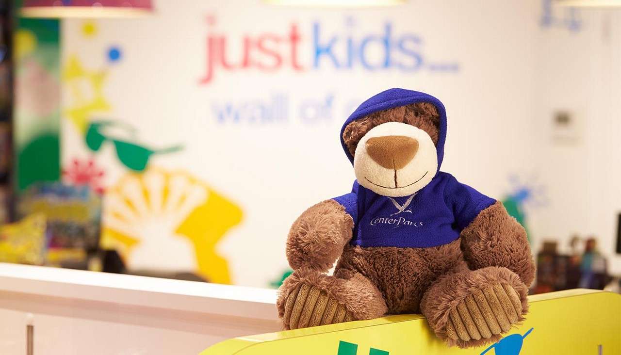 A plush teddy bear wears a blue "CenterParcs" hoodie and sits on a counter; in a colorful children’s area, blurred wall text reads: "justkids.... wall of".
