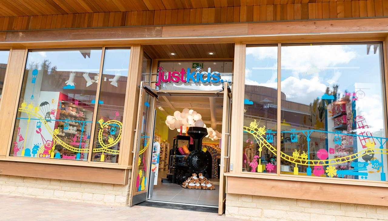 Storefront welcomes shoppers, open glass doors revealing a black train display and plush bears; bright decals decorate large windows. Context: wood-framed building, playful interior displays. Text: justkids...