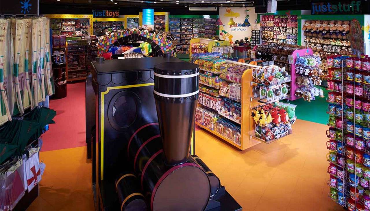 Toy train display stands at the front, while customers would walk through colorful aisles. Context: bright toy store packed with plush animals, candies, and boxed toys on racks. Text: justtoys; justkids wall of cool; juststuff.