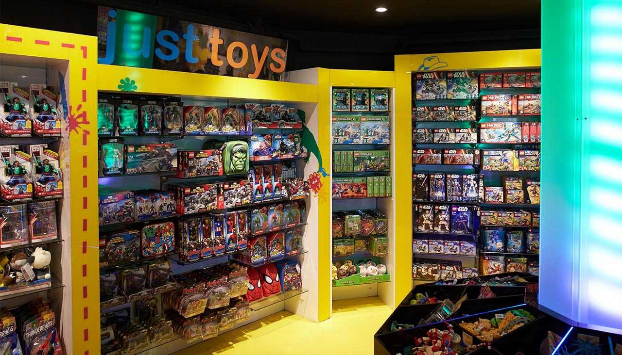 Toy shelves display action figures and building sets in a brightly lit store with yellow framing and neon glow. Visible text: just toys, LEGO, STAR WARS, NINJAGO, MINECRAFT, Thunderbirds, Avengers.