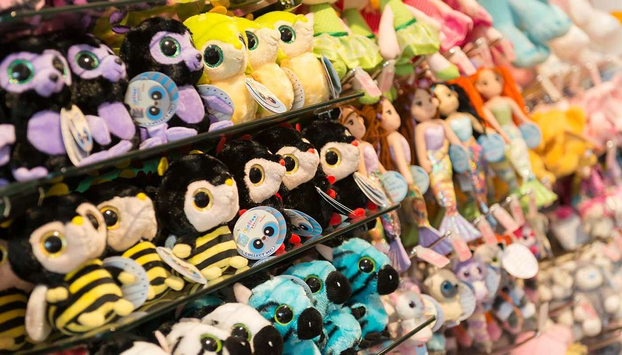 Plush toys sit arranged on metal shelves, displayed for sale; bee, penguin, lemur, and blue animal plushies line up while mermaid dolls hang nearby in a toy store. Tags read: "COLLECT ME!"