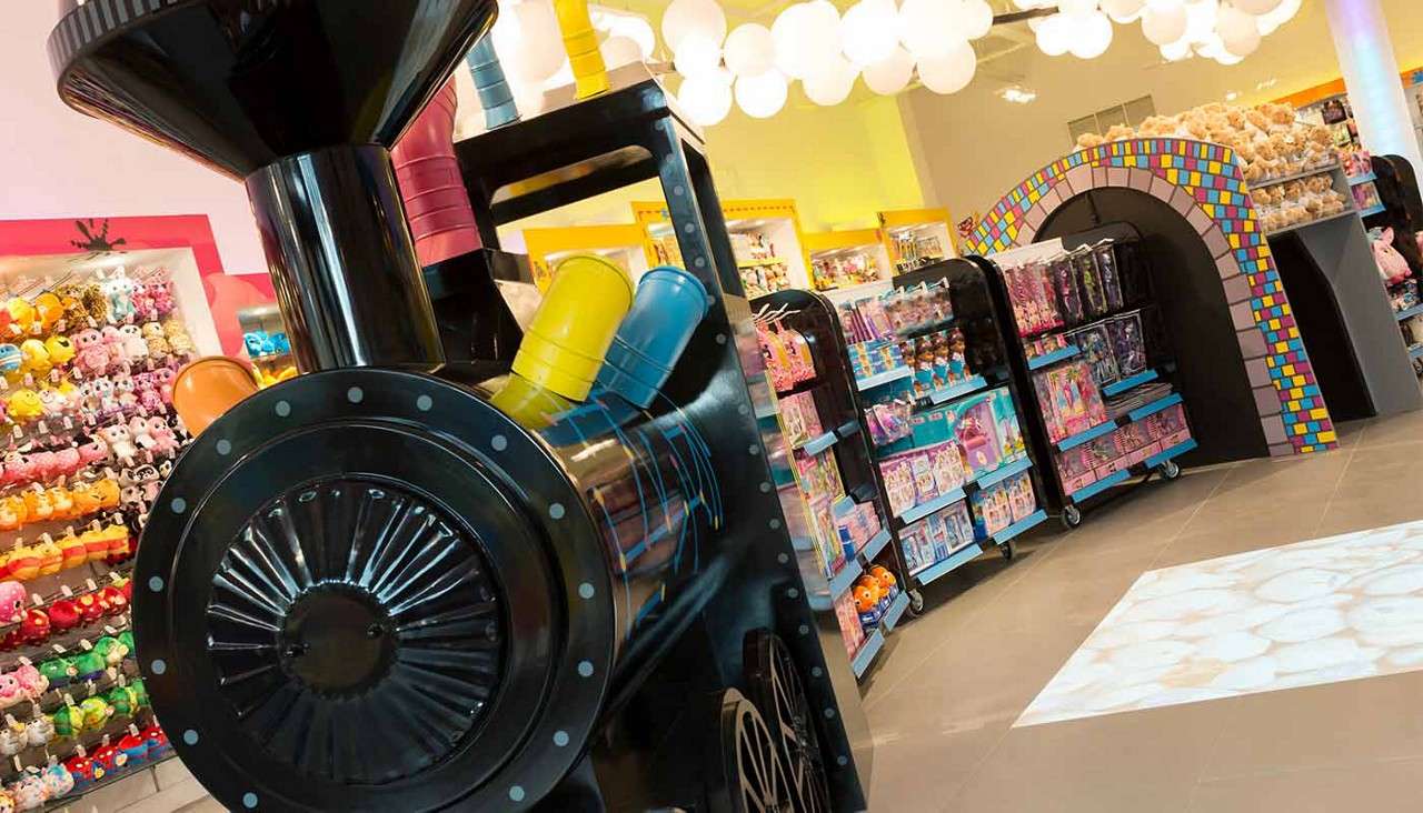 Large black toy-train display anchors foreground, facing aisles; shelves overflow with plush animals and dolls under bright round lights, beside a colorful tiled tunnel arch in a cheerful toy store.