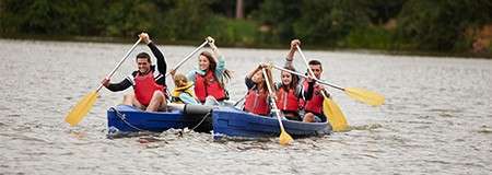 Paddlers stroke yellow-bladed oars in unison, six people wearing red life vests, traveling in a wide blue canoe across a calm lake with a tree-lined shore.