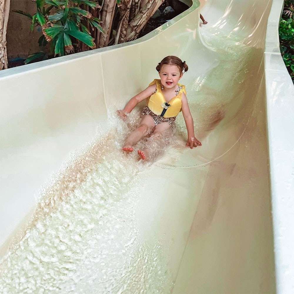 Child wearing a yellow life jacket slides feet-first down a pale water slide, splashing through shallow water; tropical plants line the slide’s edge in a waterpark setting.