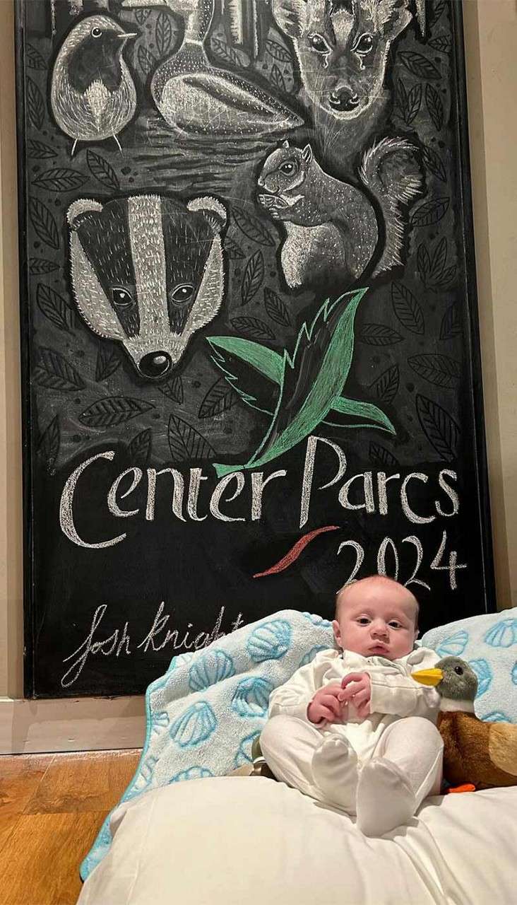 Baby sits on a cushion, holding a duck plush, in front of a chalkboard covered with woodland animal drawings. Text: “Center Parcs 2024” and “josh knight”.