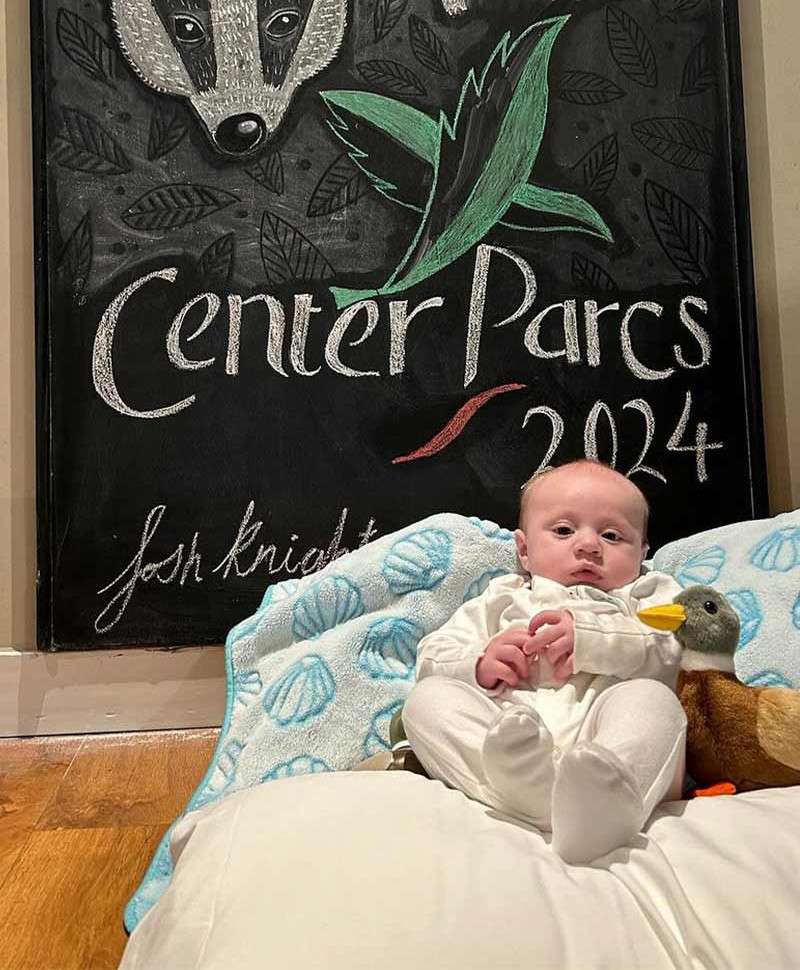 Baby sits propped on cushions, holding a plush duck; behind, a chalkboard with a badger drawing and leaves. Text: “Center Parcs 2024” and “josh knight.”
