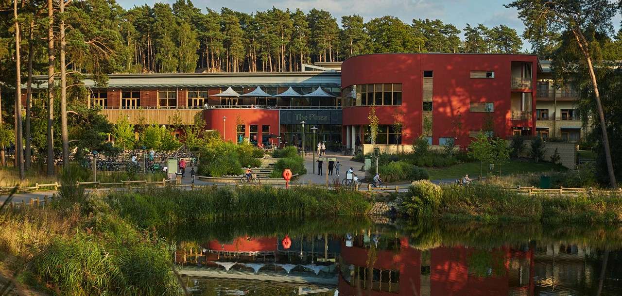 Curved red building hosts visitors walking and cycling along paths, reflected in a small lake; forested trees and landscaped gardens surround it. Visible sign: The Plaza.