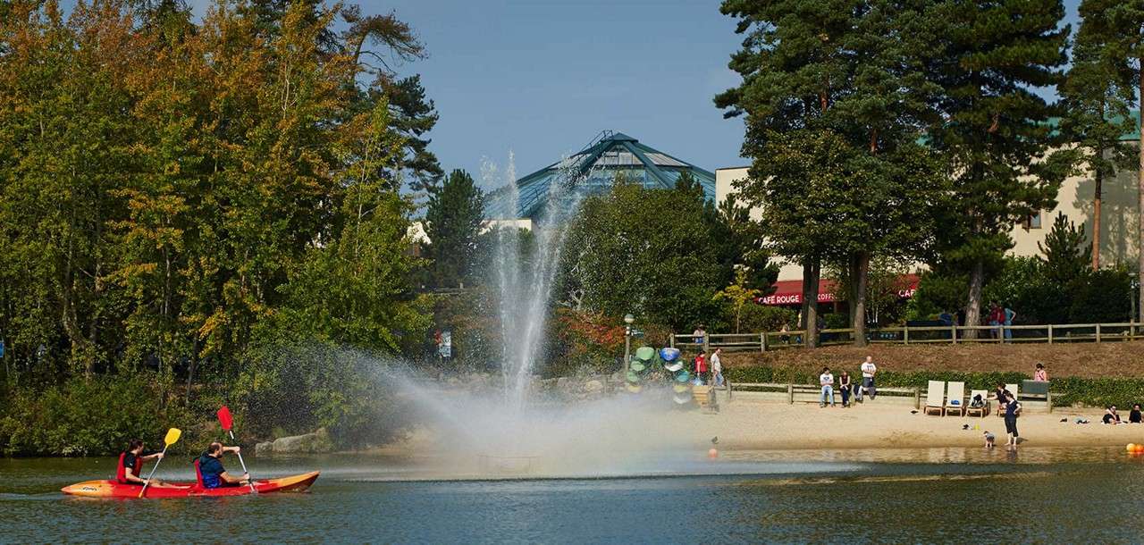 Kayakers paddle a red canoe across a lake as a fountain sprays; nearby, people relax on a sandy beach before trees and buildings with awnings reading CAFE ROUGE, CAFE ROUGE.