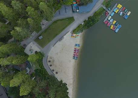 Colorful paddle boats sit moored on calm lake water, beside an empty sandy beach; pedestrians walk along curving paved paths bordering a grassy area and dense trees, viewed from above.