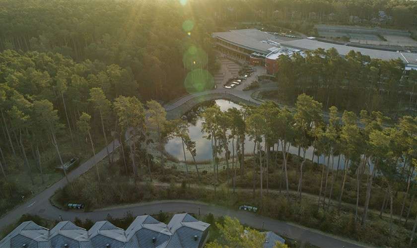 Tree-lined pond reflects sunlight, bordered by curving roads and pathways, while a large low-rise complex and parking sit nearby; dense pine forest surrounds the area under a low, hazy sun.