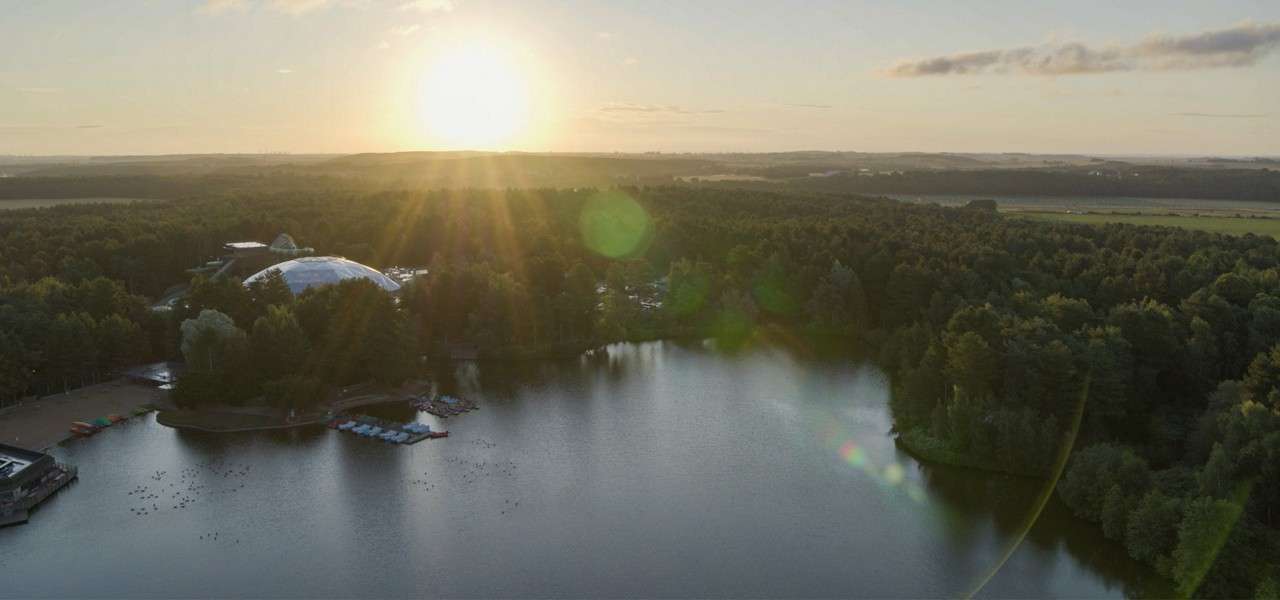 Sun rises, casting rays over a calm lake; water reflects light. Surrounding forest encloses a shoreline with docks and small boats; a large dome sits among trees, beyond fields.
