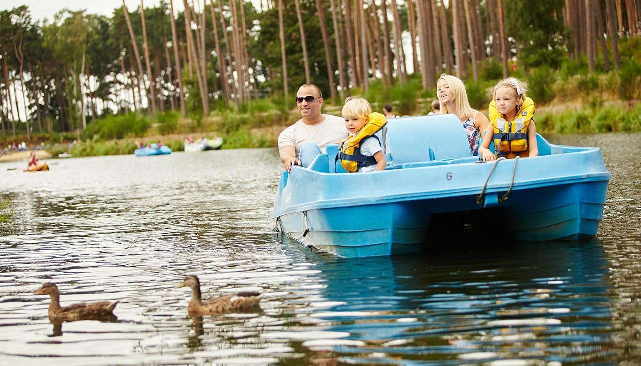 Two adults and two children in a blue pedal boat watch two ducks ahead, pedaling on a calm lake amid tall pines, with other boats behind. Visible text: 6.