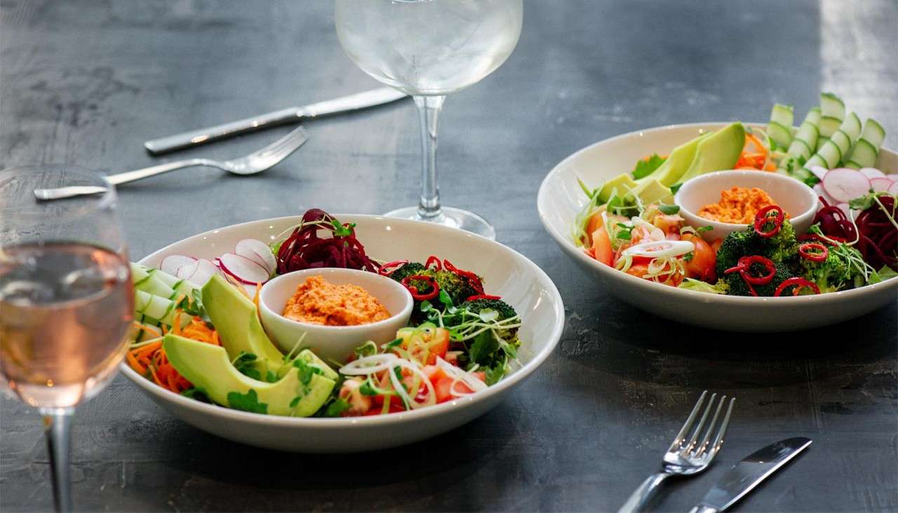 Two salad bowls rest on a table, displaying avocado, cucumber, radish, shredded carrots, beet, broccoli with chili, and a central orange dip; wine glasses and cutlery create a dining context.