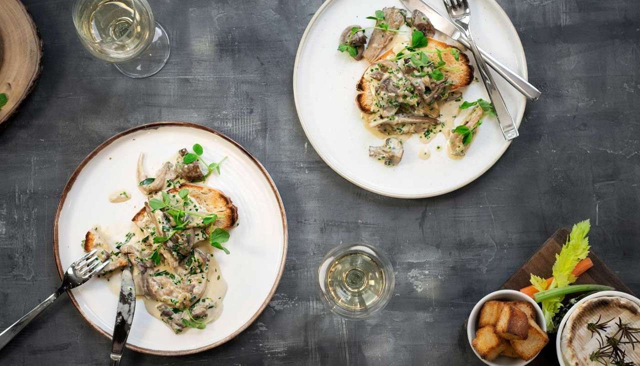 Mushroom toast sits under creamy sauce, garnished with herbs; forks rest nearby. Two plates share the table with white wine glasses, a bowl of croutons, and vegetable sticks on a gray surface.