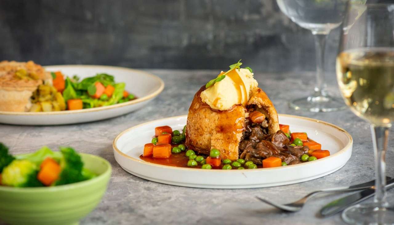 Meat pie topped with mashed potato sits cut open, spilling stew and gravy, ringed by peas and carrots, on a table with salad plate, bowl, wine glass, and cutlery.