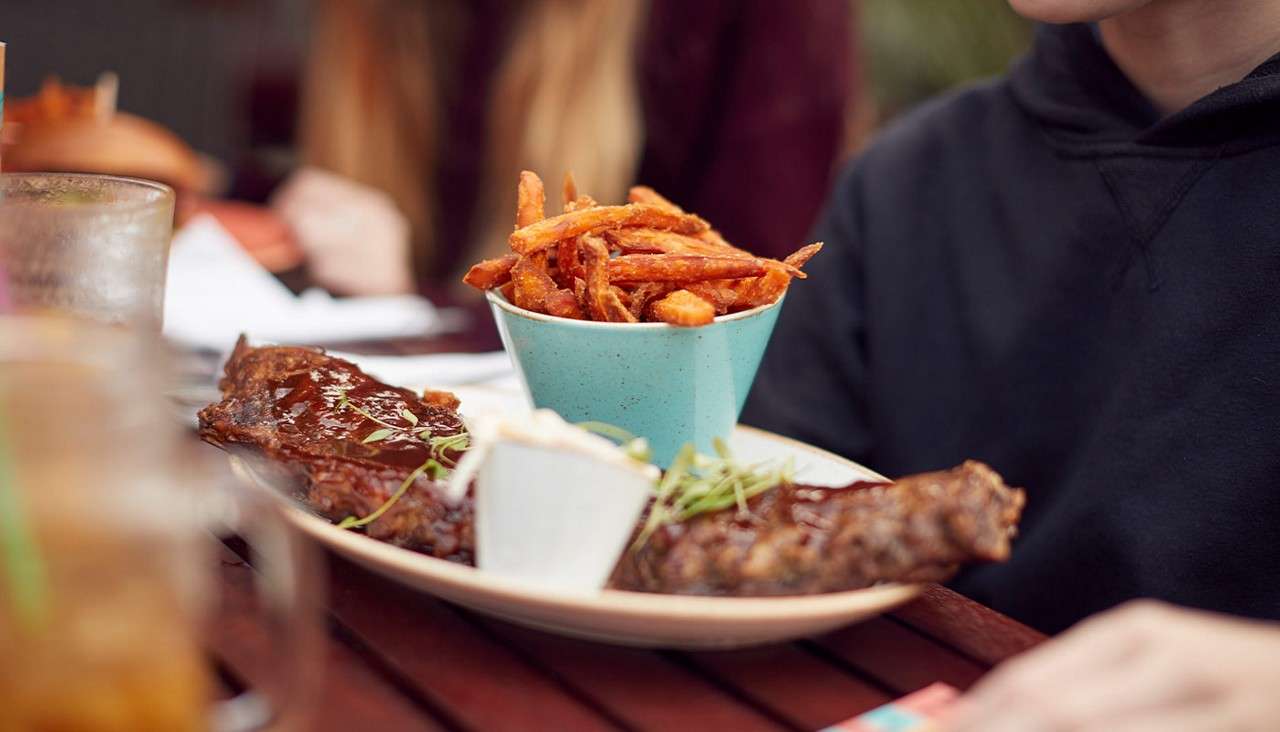 Barbecue ribs sit glazed on a plate, with a cup of sweet potato fries and a sauce dish, as diners gather around a wooden table in a casual background.
