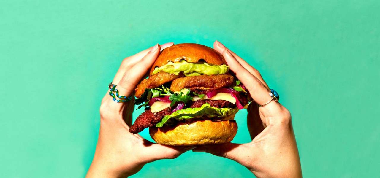 A stacked burger is held with both hands, showcasing glossy bun, green spread, crispy patties, onion rings, lettuce, cheese, and pickled onions against a plain teal background.
