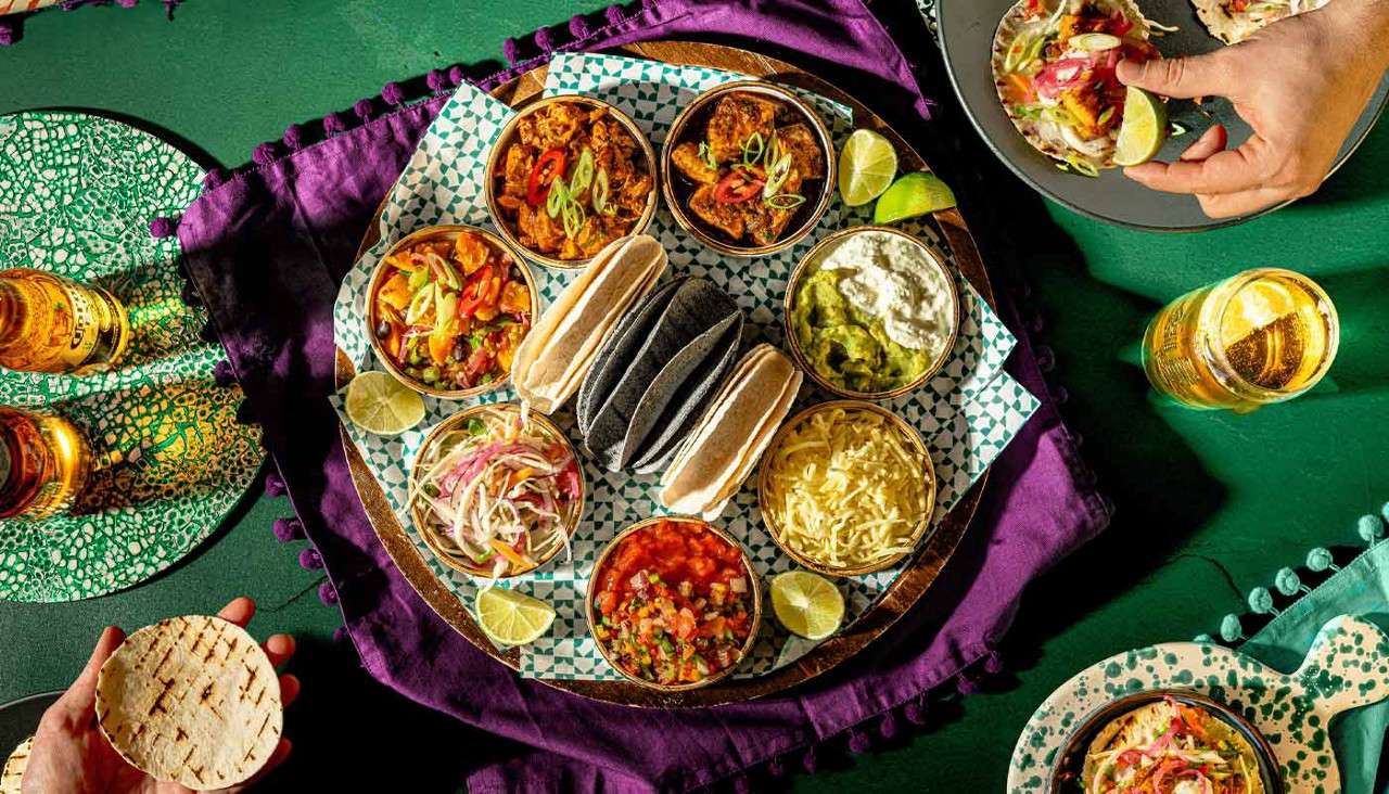 Assorted taco fillings sit in small bowls, with tortillas stacked upright, while hands assemble tacos; lime wedges and beers surround the platter on a green table with a purple cloth.