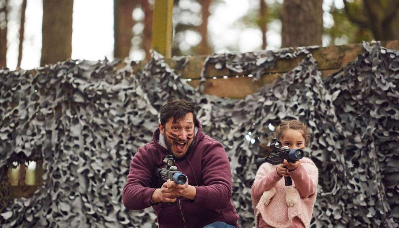 Man and young girl aim toy laser guns while crouching and smiling, faces painted, in a woodland play zone with camouflage netting.