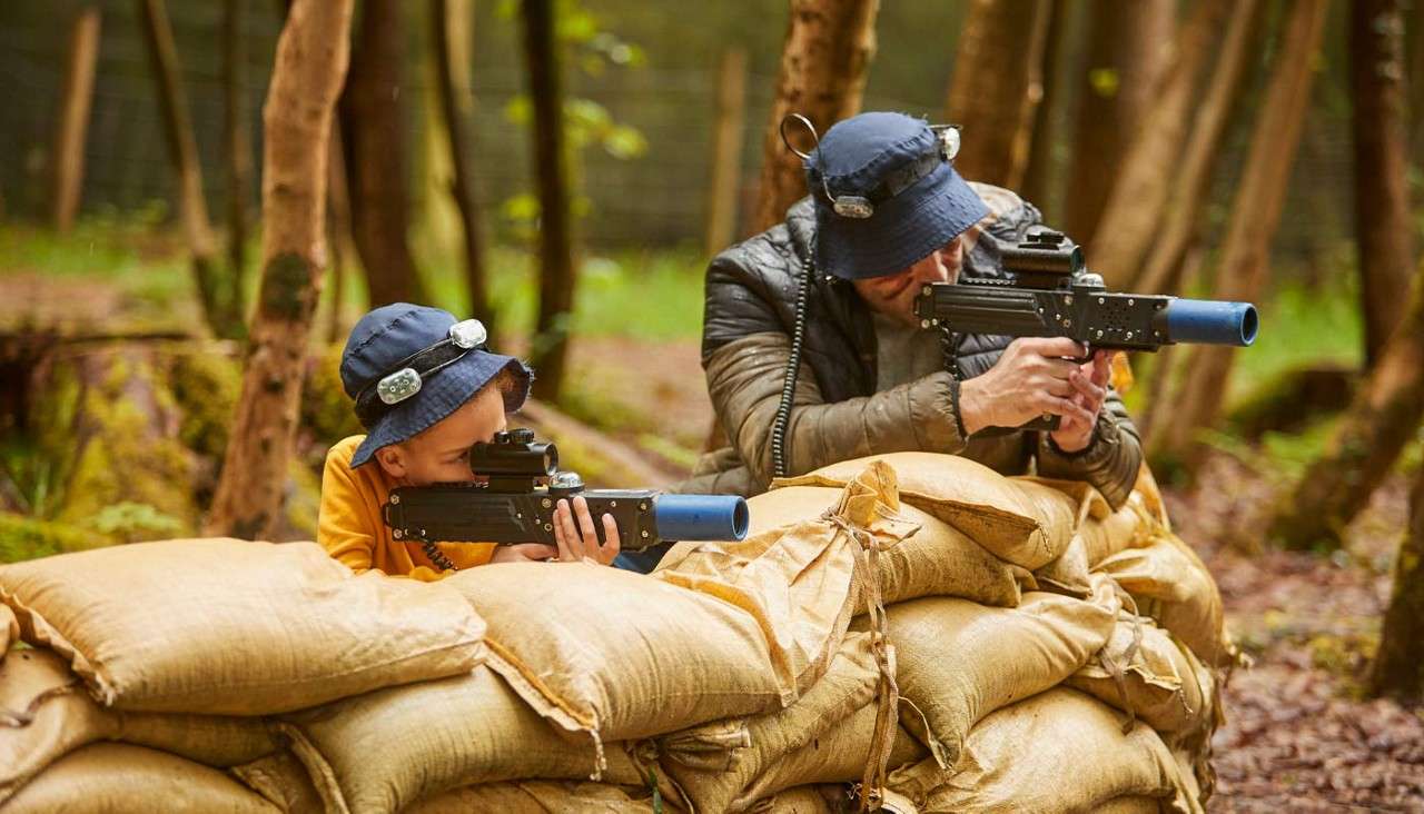 Two players aim laser-tag rifles, crouching behind stacked sandbags, wearing sensor hats and headsets, in a wooded outdoor arena with trees and soft, brown leaf-covered ground.