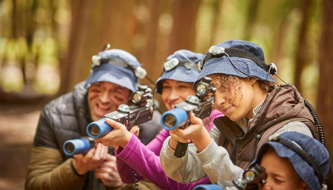 Group of players aim laser tag blasters, focusing down sights. They wear bucket hats with sensors and vests. Context: wooded outdoor course with soft, blurred trees.