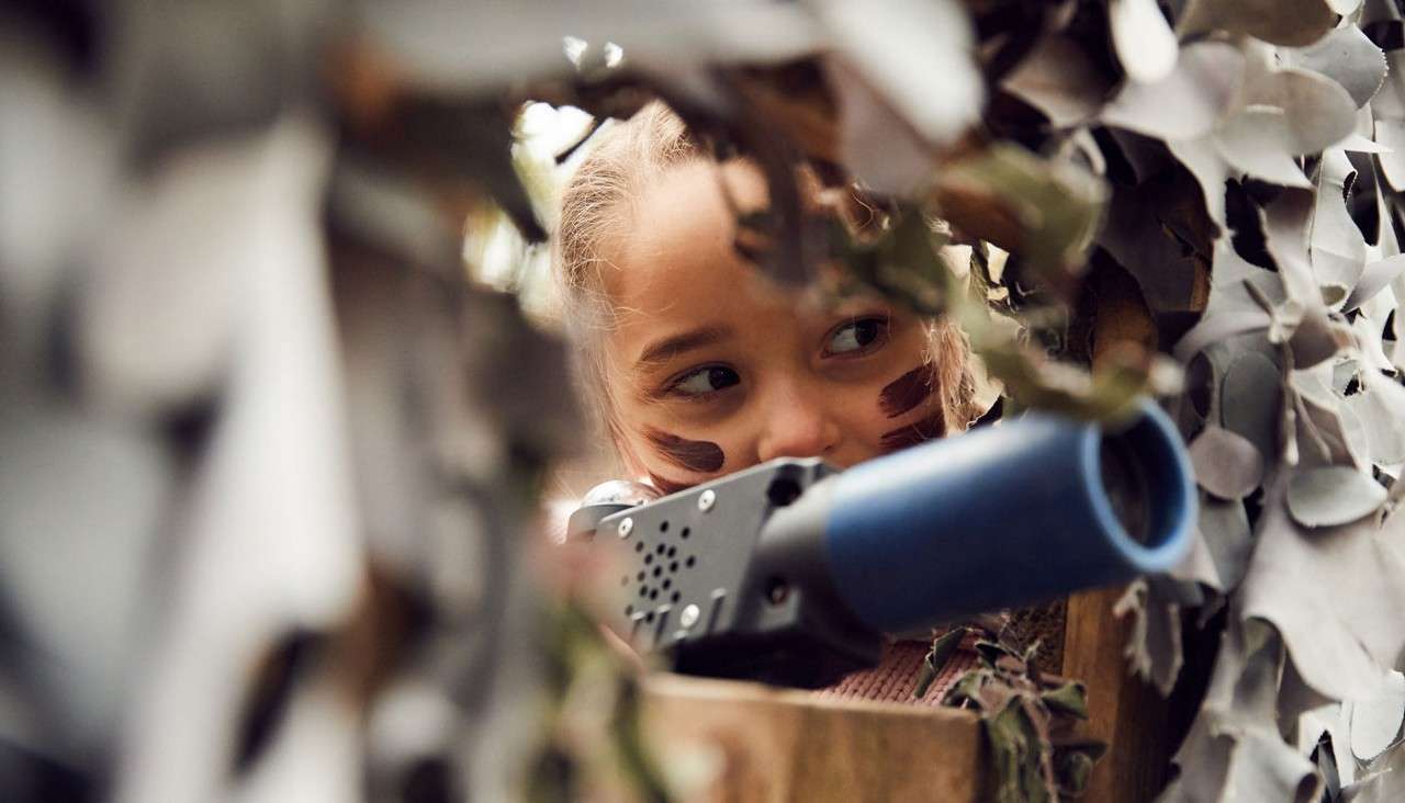 Child with face paint aims a plastic blaster, peering cautiously through gaps in camouflage netting from a wooden hide or fort during play.