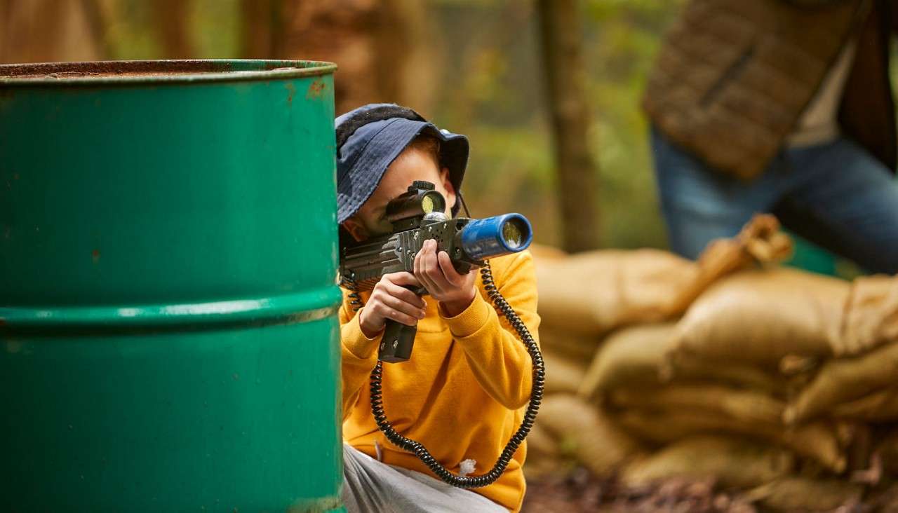 Child in yellow sweater aims a laser-tag gun from behind a green barrel, crouching outdoors. Coiled cable connects gear; sandbags and a standing adult appear in the wooded background.