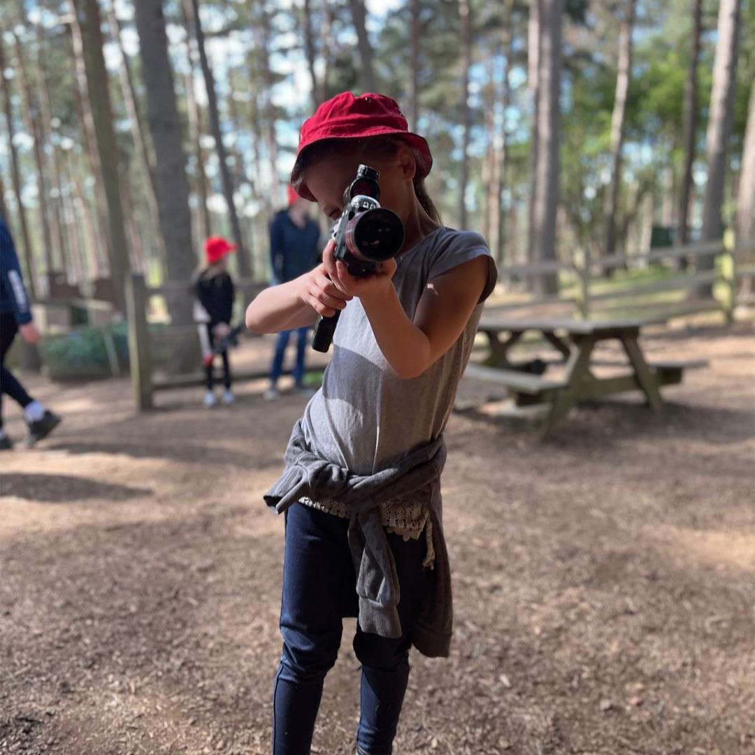 Child in a red bucket hat aims a laser-tag gun forward, standing on dirt in a wooded park with picnic tables and a fence; other people linger blurred in background.